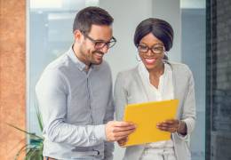 Two colleagues, a man and a woman, share a moment of collaboration as they review a yellow folder together. Both are dressed in business casual attire and appear engaged and cheerful, suggesting a positive and productive work environment. The background includes modern office elements, emphasizing a professional setting.