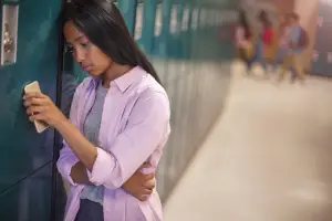 A sad student stands by lockers in school holding a smartphone