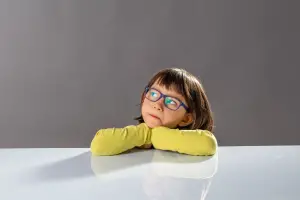 A young child with short brown hair and blue glasses rests their chin on their hands, looking thoughtfully to the side. Wearing a bright yellow long-sleeve shirt, the child is set against a plain gray background, creating a calm and contemplative atmosphere