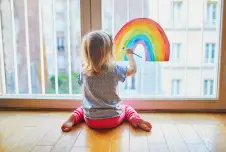 A young girl sitting on the floor in front of a large window, painting a rainbow on the glass